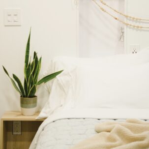 The image frames the left side of a queen bed with crisp white linens, a soft blue comforter and a rumpled peach throw blanket against a white wall with a wood beaded garland hanging above. A simple bedside table is adorned with a lush green snake plant.