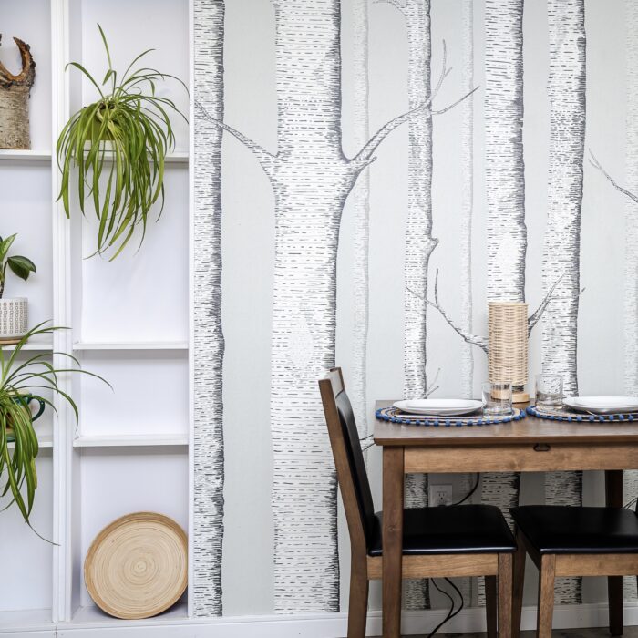 A square table is set for two in front of an accent wall papered in a soft green birch tree motif. A white inset shelf beside the accent wall is filled with wood pieces and lush green potted plants.