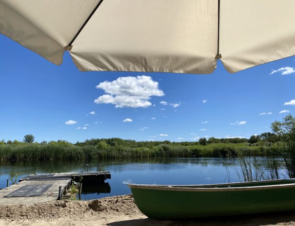 Looking out from under a tan sun umbrella, a green canoe sits on a sandy beach in front of a small blue lake with a wooden dock floating against the shore. Green cattails, gentle hills & a row of trees in the distance line the far shore.