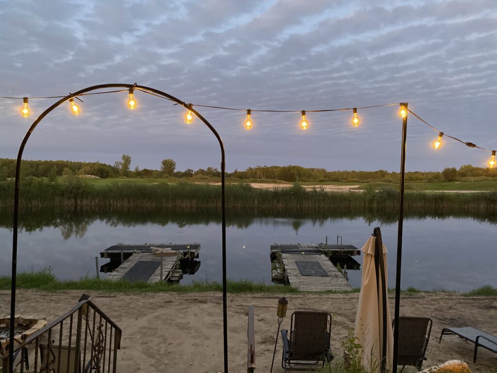 A string of lights is lit up in the dusk against an evening sky streaked with purple clouds. They frame a view of a sandy beach with two docks floating on a small lake reflecting the purple sky. Gentle green hills of a golf course can be seen on the opposite shore. In the foreground, a closed sun umbrella stands tall over reclining chairs on the beach.