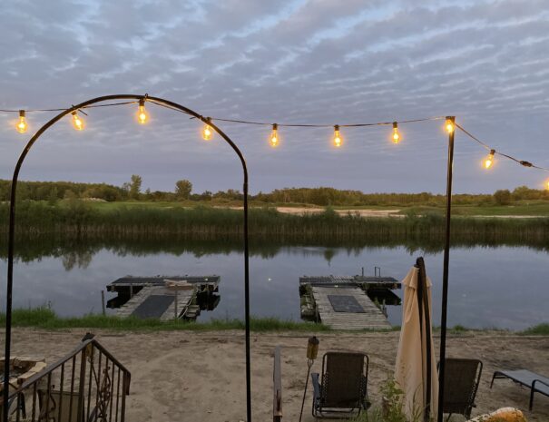 A string of lights is lit up in the dusk against an evening sky streaked with purple clouds. They frame a view of a sandy beach with two docks floating on a small lake reflecting the purple sky. Gentle green hills of a golf course can be seen on the opposite shore. In the foreground, a closed sun umbrella stands tall over reclining chairs on the beach.