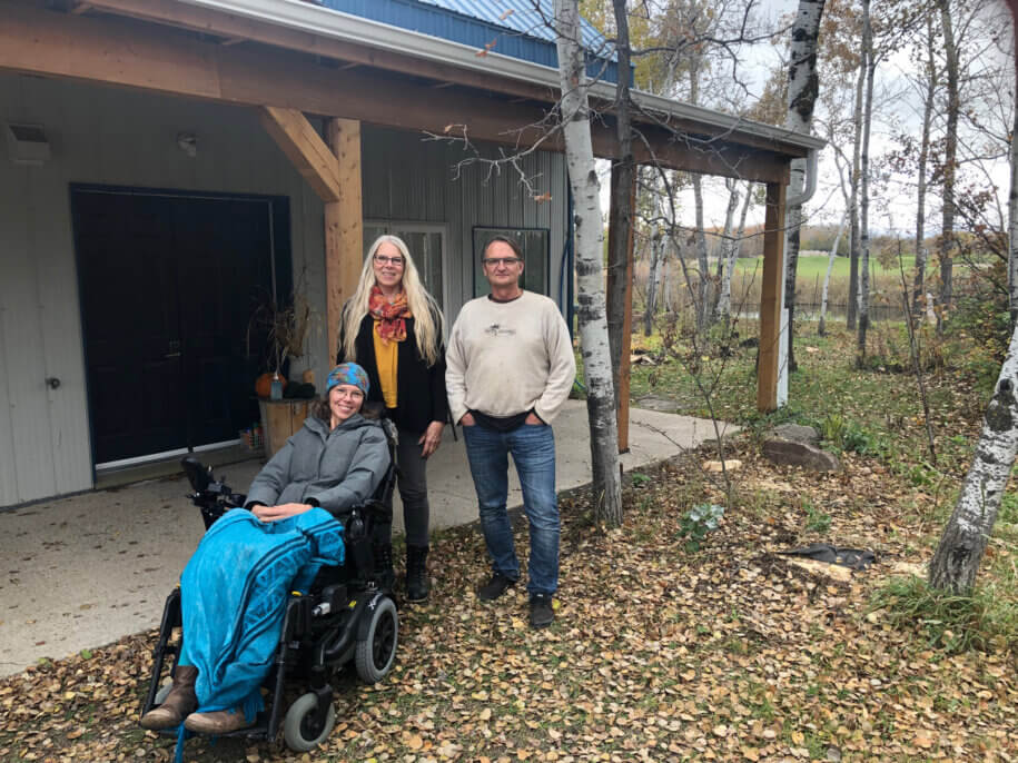 A woman in a wheelchair with a bright blue lap blanket poses with a woman with long blonde hair and a man with dark hair in front of a grey building with a warm wood post veranda and a blue roof. The ground is carpeted with fallen autumn leaves.