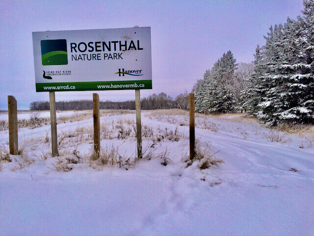 A large white and green municipal sign mounted on wooden supports reads ‘Rosenthal Nature Park’. A walking trail has been worn into the snowy landscape, heading through frozen grasses towards a treeline in the distance.