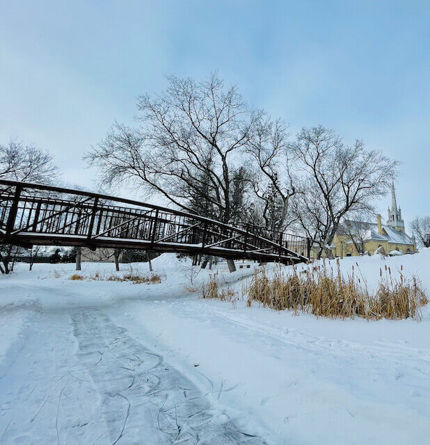 A skating trail is cleared in the snow on a winding frozen river. A trail for walking has been worn into the snow beside the skating trail. A walking bridge arches gently over the river, with several large bare trees silhouetted black against the blue sky. A yellow church with a tall white steeple peeks through the trees on the right hand side.