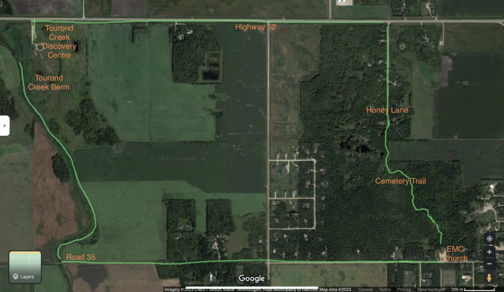 A top-down satellite image of 2 square miles of roads framing rural countryside. A green hand-drawn line sketches out a walking path along the major geographical features of the photo.